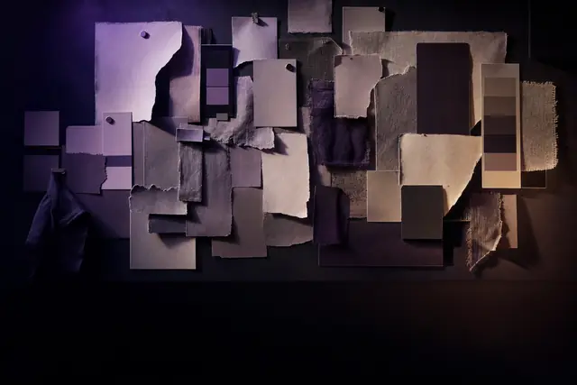 A wall of pinned blank paper swatches and fabric samples in a dark studio, raked by warm and violet light.
