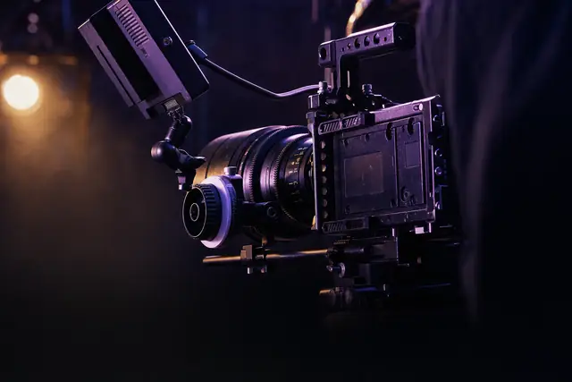 A close view of a cinema camera lens and monitor arm in a dark studio, edged with violet light.
